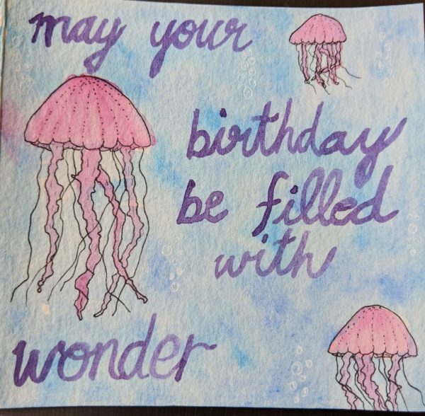 3 pink jellyfish float in the blue ocean. Purple watercolor letters say "may your birthday be filled with wonder"