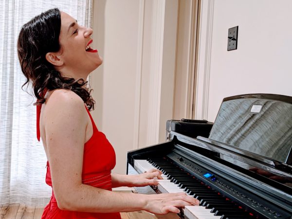 Angelle Conant sits at a piano in a red halter dress, singing and playing the piano. 