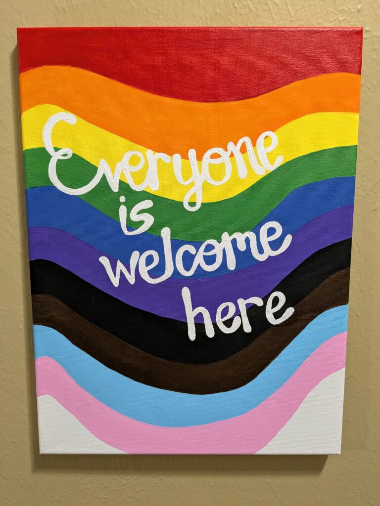 A canvas painting with red, orange, yellow, green, blue, purple, black, brown, baby blue, baby pink, and white waves. On top of the colorful waves in white lettering are the words "Everyone is welcome here."